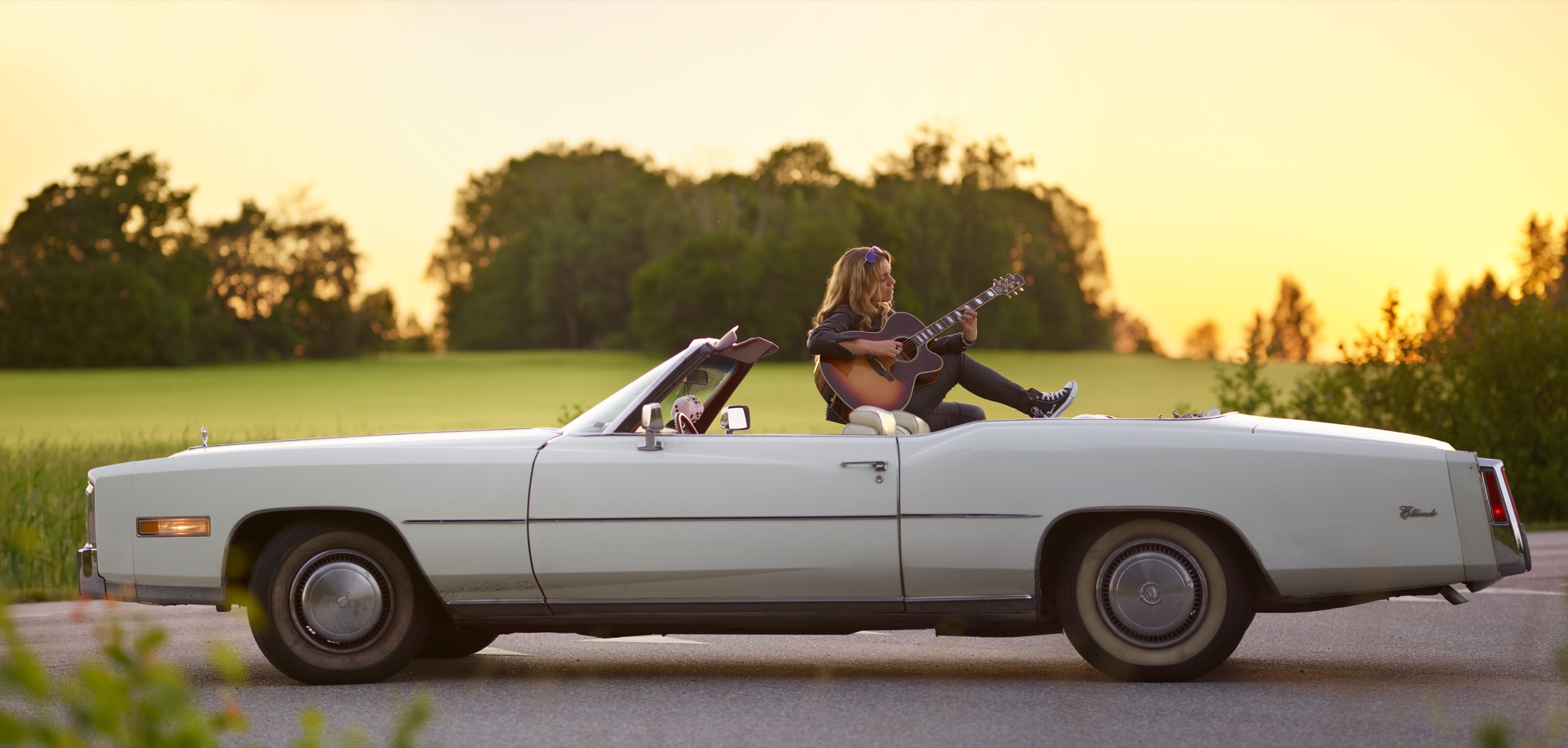 Alexandra Jardvall spelar gitarr sittande på en vit Cadillac Eldorado cabriolet vid ett fält i solnedgången. Foto: Irsta Foto
